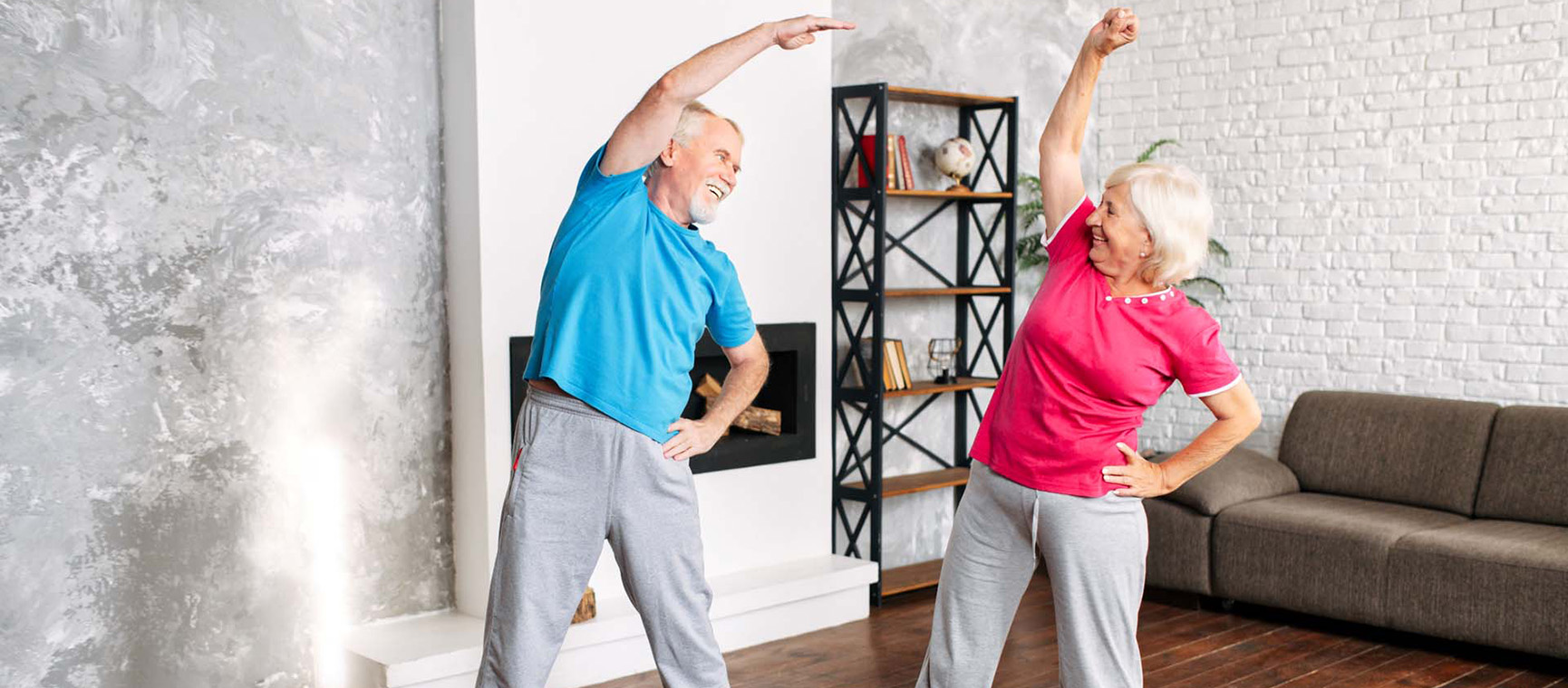 A man and woman standing next to each other in a living room, dressed in t-shirt and tracksuit bottoms doing side stretches