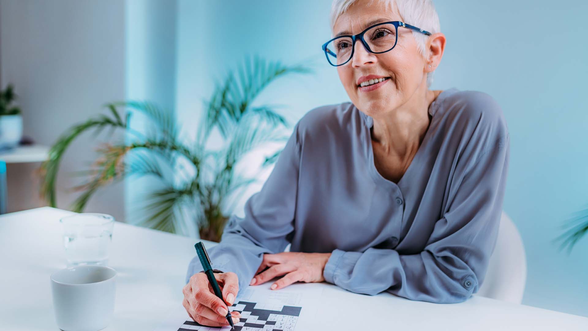 Silver-haired older lady with short hair wearing blue glasses and smiling while doing a crossword