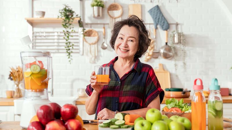 Woman drinking fresh juice she has just made in her kitchen. She has fruit in front of her and a juicer to the right of her.