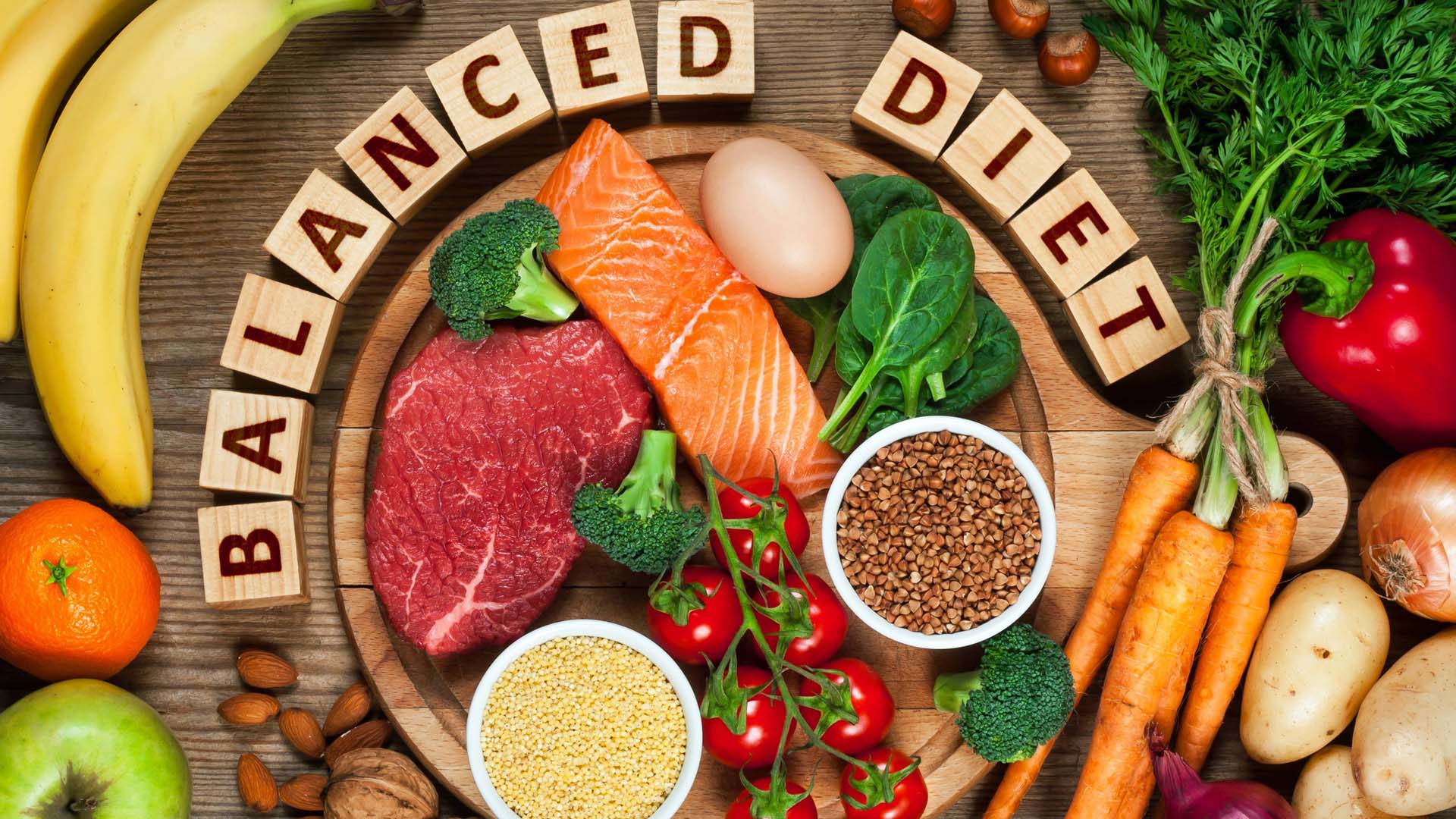 A flat lay image of fruit, vegetables, meat, fish and dairy products displayed on a wooden surface, with the words 'balanced diet' spelled out in wooden letters