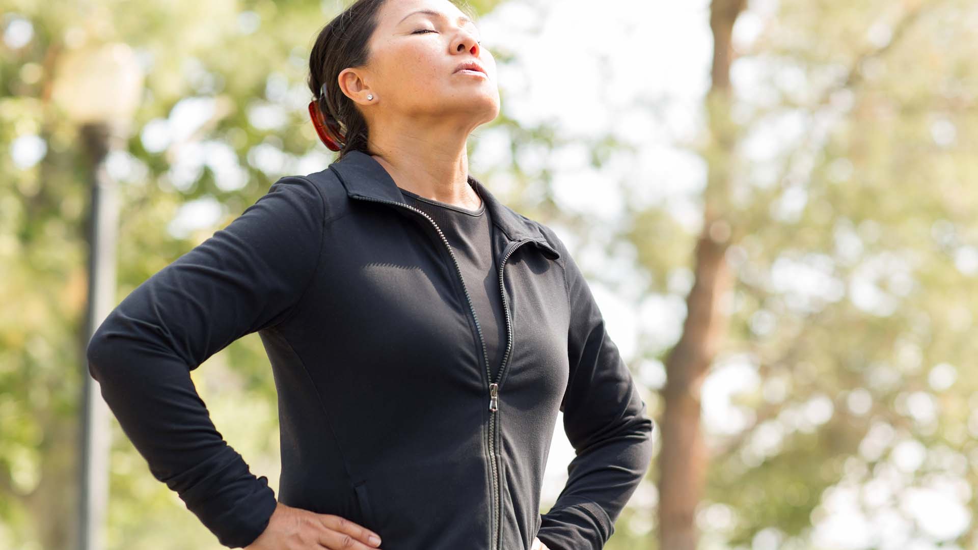 A woman in a dark top taking a deep breath outdoors.