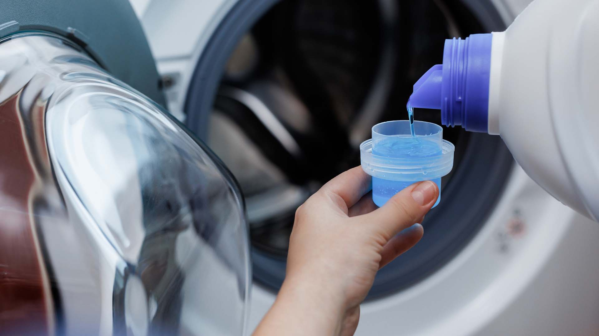 Close-up of liquid washing detergent being poured into a cup with a washing machine in the background
