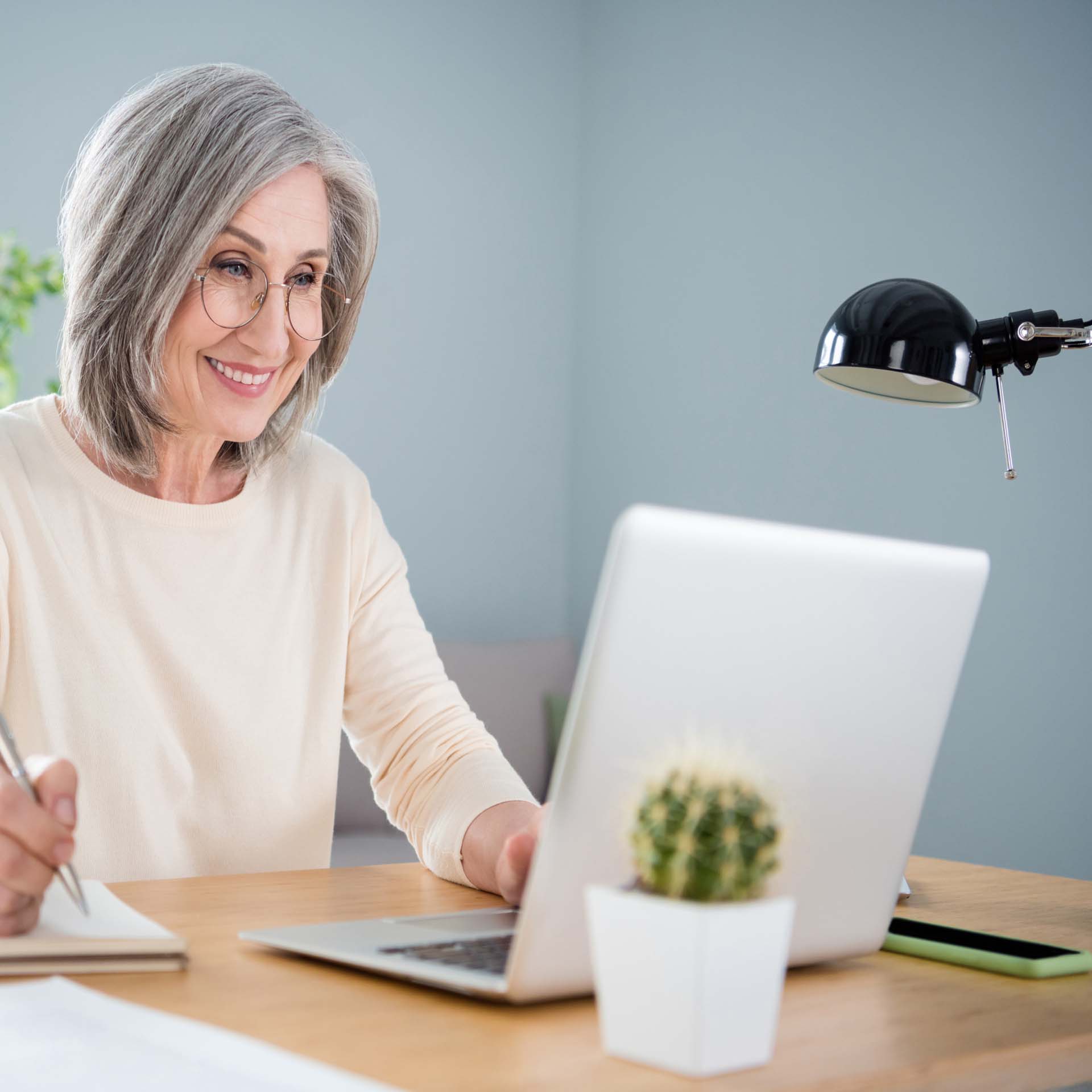 A stylish mature woman with a grey bob and glasses sitting at a desk using a laptop