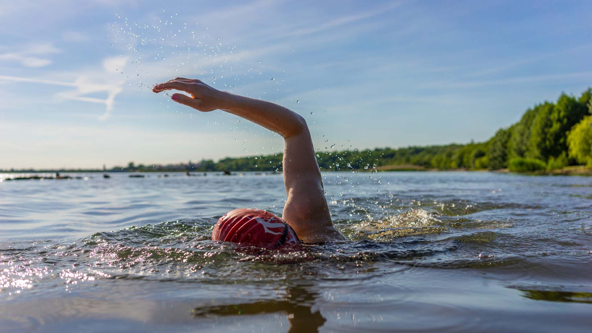 Woman swimming in a lake. While her face is concealed, she has one arm raised and is wearing a red swimming cap.