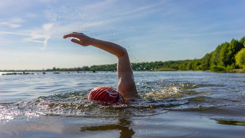 Woman swimming in a lake. While her face is concealed, she has one arm raised and is wearing a red swimming cap.