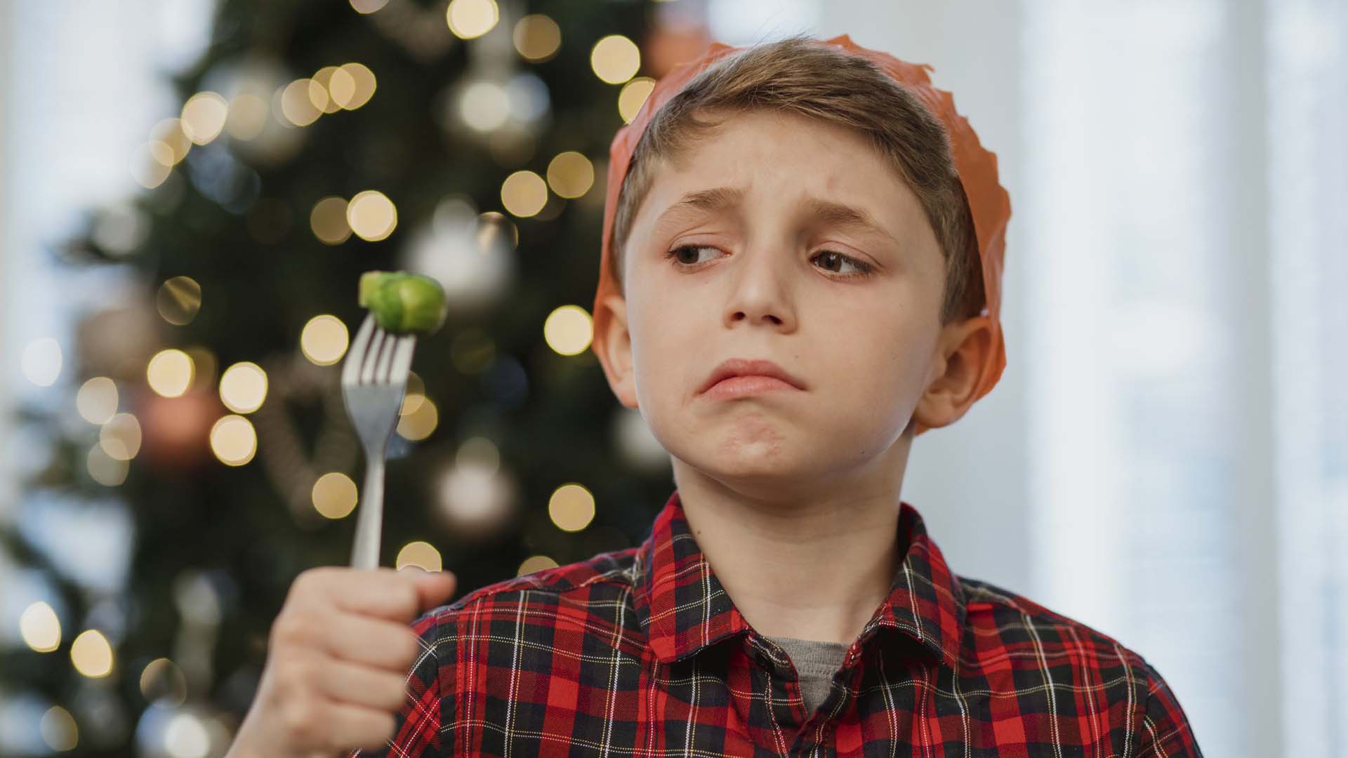 A child wearing a Christmas cracker hat looking suspiciously at a Brussels sprout speared on the end of a fork