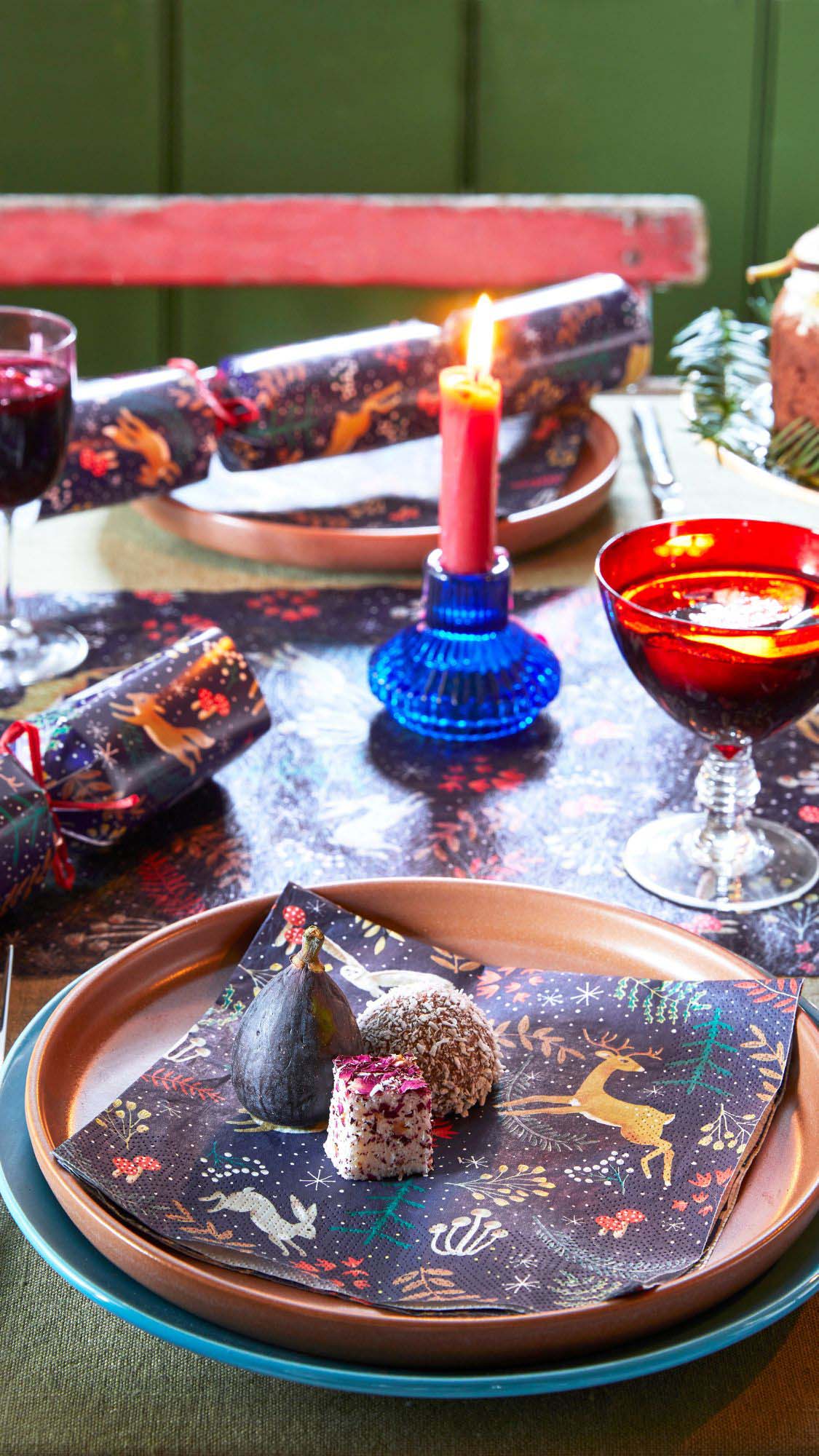 A festive table setting with matching napkins, cracker and table runner, a red candle in a blue candle holder and bright wine glasses