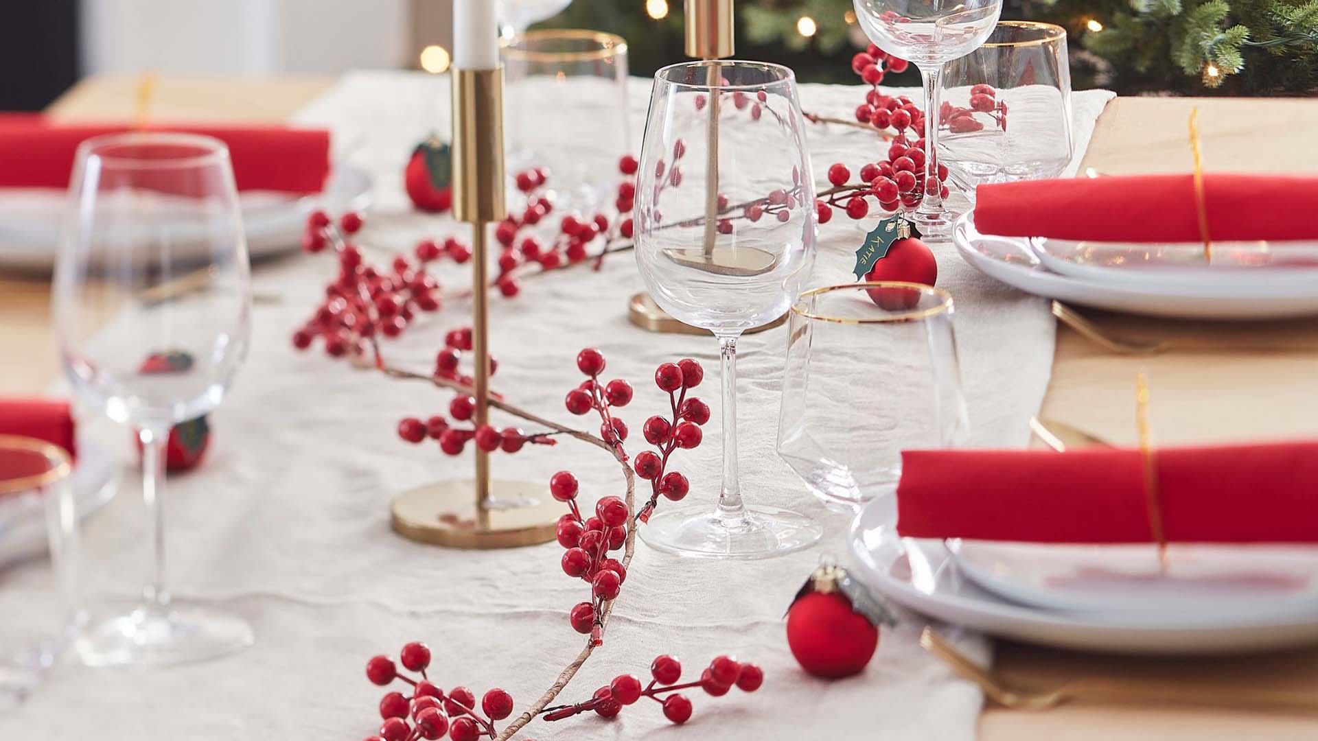 A festive table setting with red accents, including a branch of berries as a table runner and red napkins on top of plates