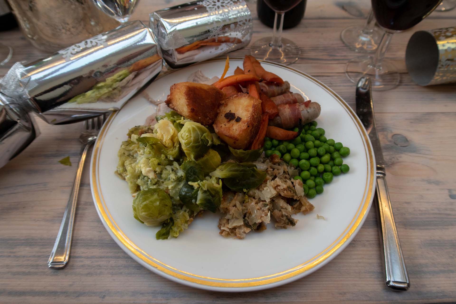 A plated Christmas dinner complete with turkey, pigs in blankets, roast potatoes, peas and Brussels sprouts, with a silver cracker next to the plate