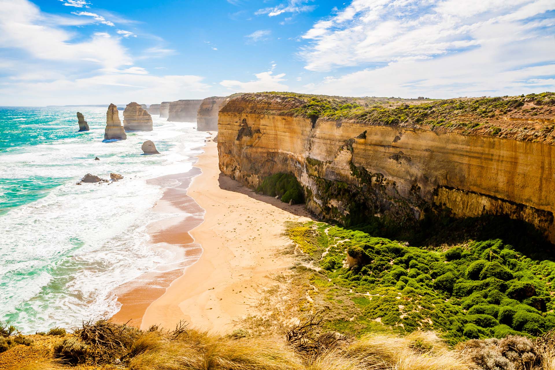 A view over cliffs above a sandy beach with rock formations out to see and waves rolling in, in Australia, with the sun shining