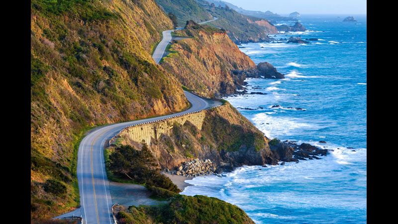 A road winding along the Pacific coast of the USA, with crashing waves to the side.
