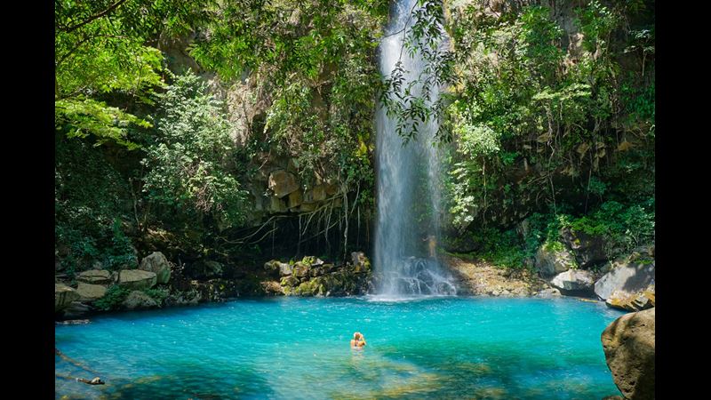 An idyllic waterfall in Costa Rica, with bright blue water and a swimmer. Surrounded by rocks and trees