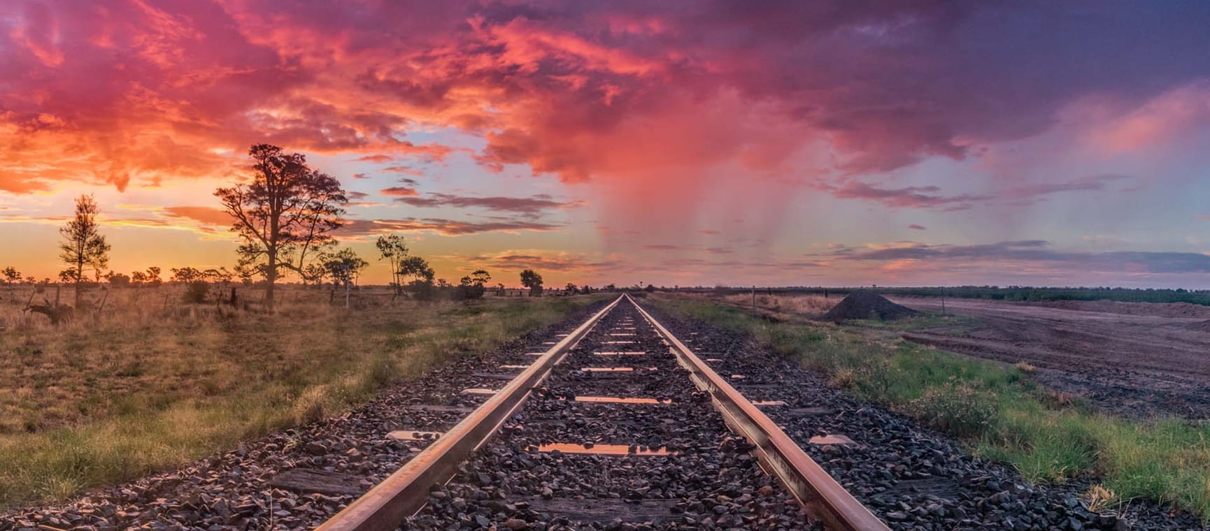 Train tracks going off into the distance across an arid landscape with a red and purple sunset.