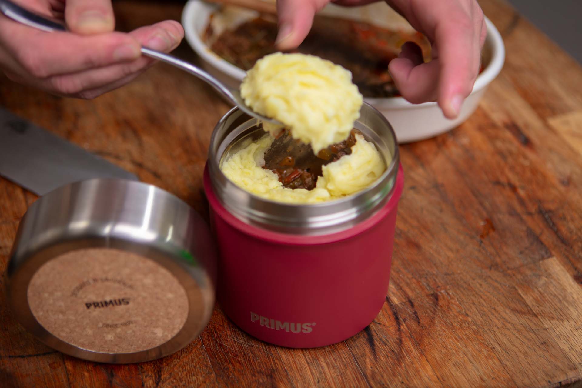 Spooning mashed potato onto the top of a shepherd's pie in a red food flask