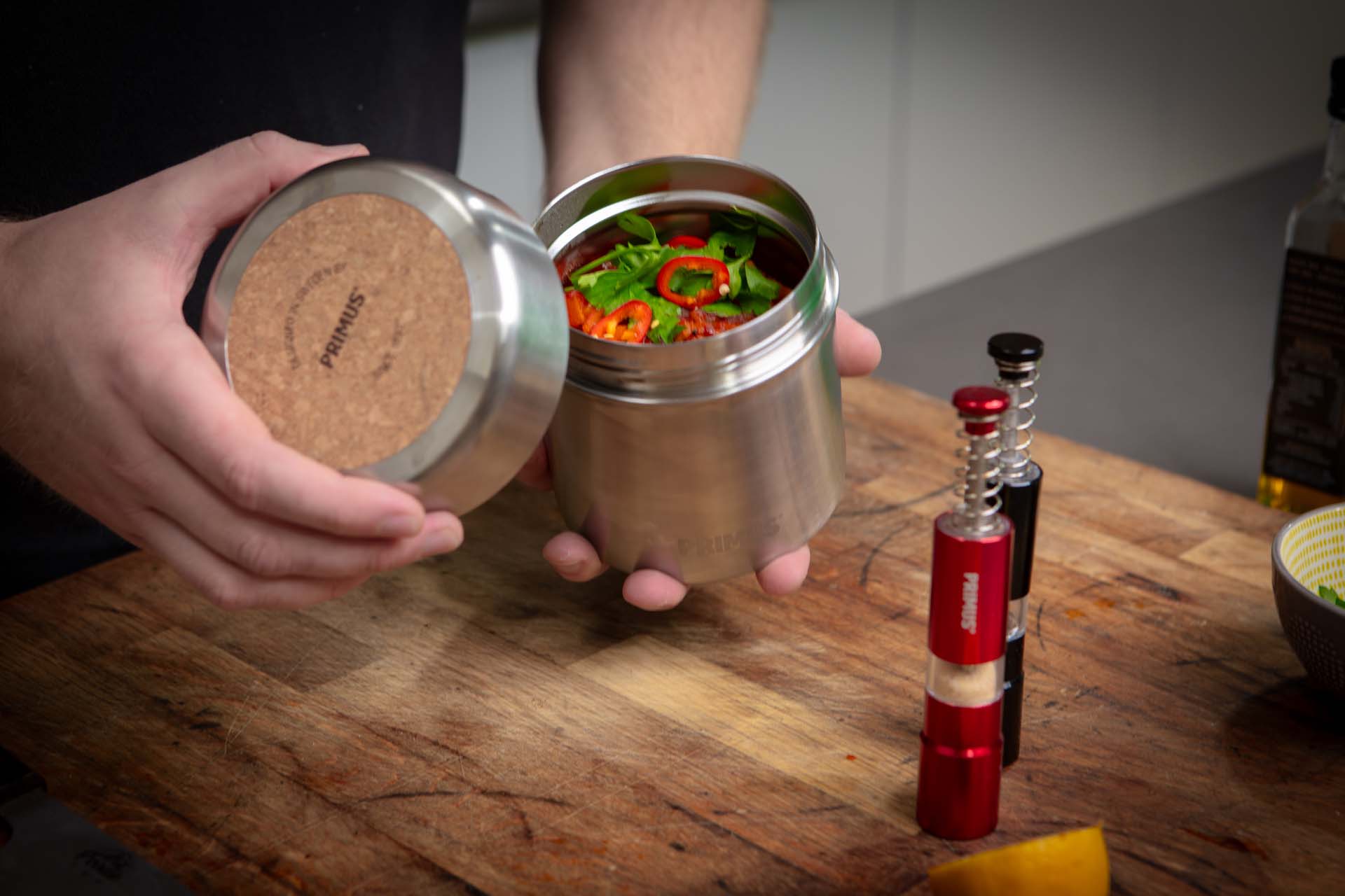 Hands holding a silver food flask containing a tagine; chopped chillies are visible on the top