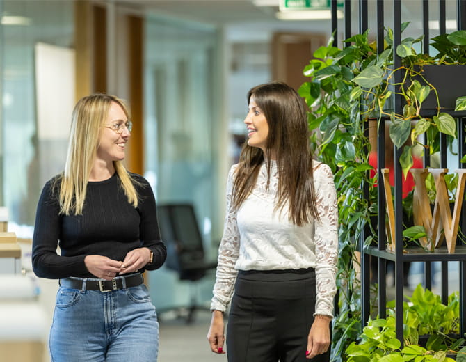 Two people walking and talking in an office with plants on the right hand side