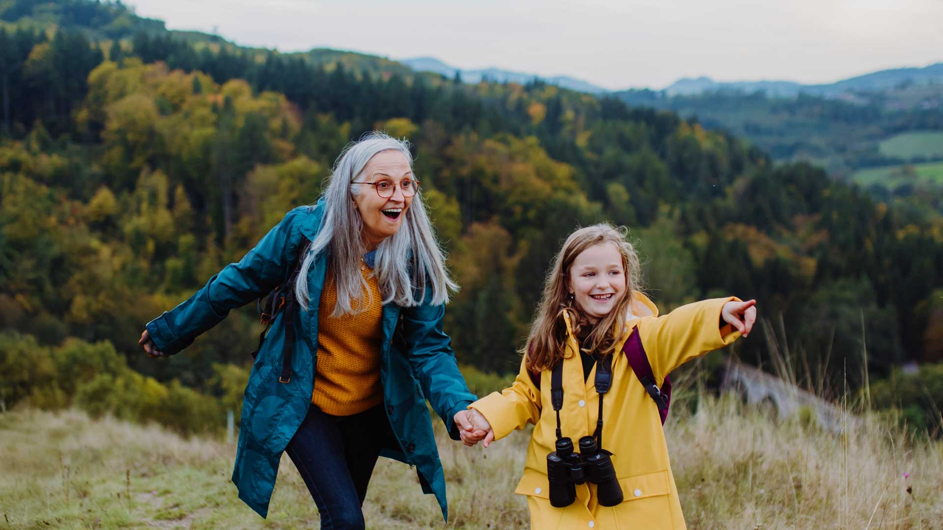 Older adult and child hiking in a grassy hill, smiling and pointing at the scenic view.
