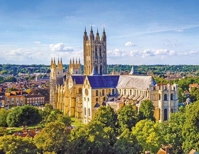 A large historic cathedral with tall towers and detailed stone architecture, surrounded by trees and a townscape under a bright blue sky.