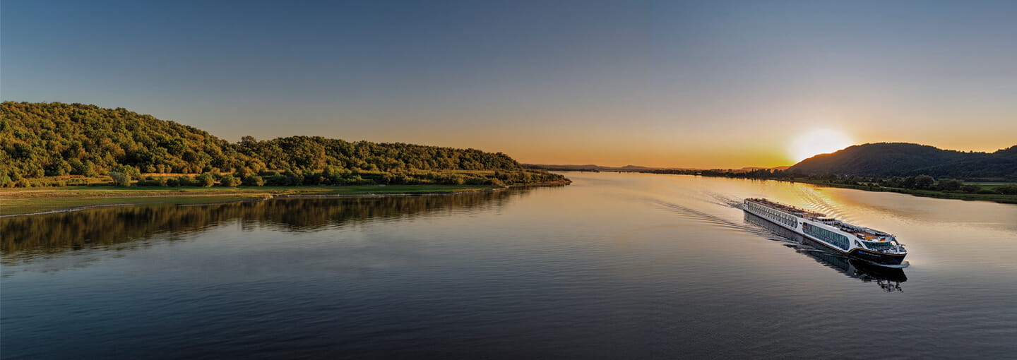 River cruise boat sailing on calm water at sunset, with hills and shoreline in the background