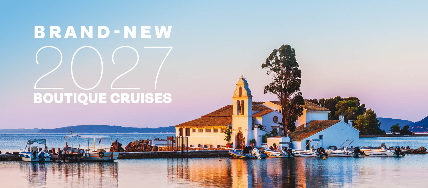 A small waterfront church and surrounding buildings on a calm shoreline at sunset, with boats docked along the pier and soft pink and blue sky reflected in the water.