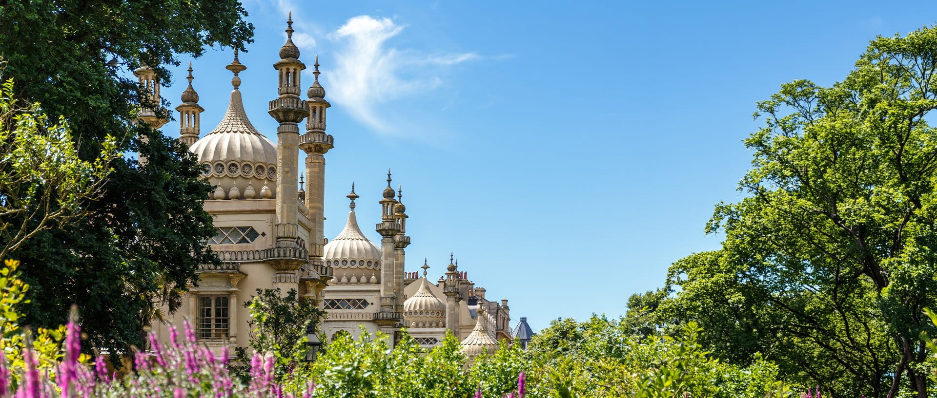 Ornate historic palace with domes and spires framed by trees and flowering gardens under a clear blue sky.