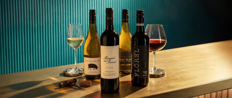 Four bottles of wine and two filled wine glasses displayed on a wooden table against a teal-paneled wall.