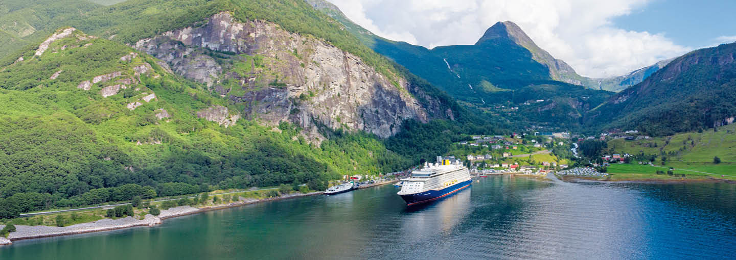 Cruise ship sailing through a fjord surrounded by mountains, green valleys, and waterside villages.