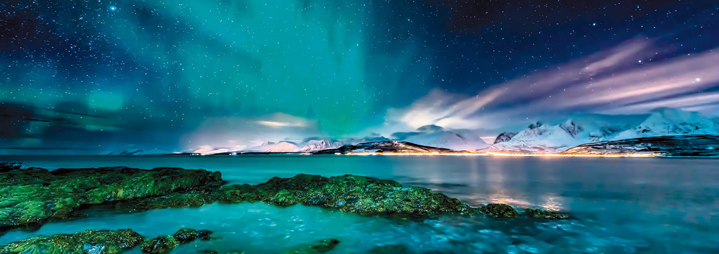 Stunning view of the Northern Lights over snow-covered mountains and a calm sea.