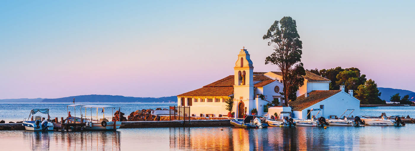 A small waterfront church and surrounding buildings on a calm shoreline at sunset, with boats docked along the pier and soft pink and blue sky reflected in the water.