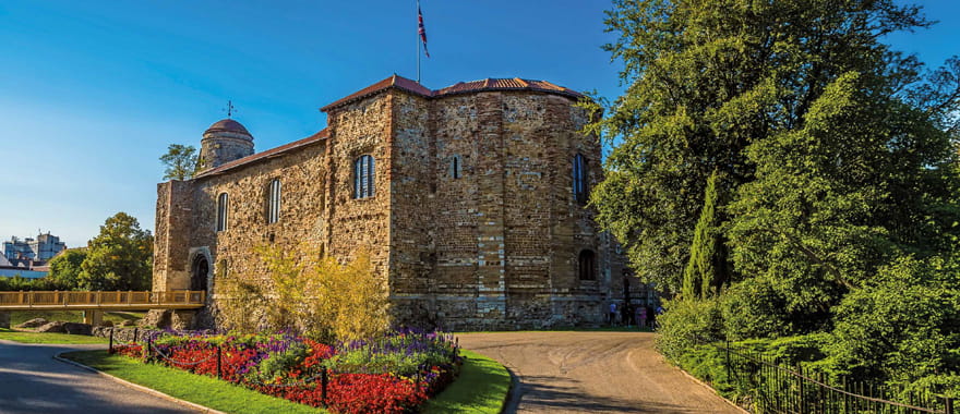 Historic stone building surrounded by gardens with colourful flowers and trees under a clear blue sky.