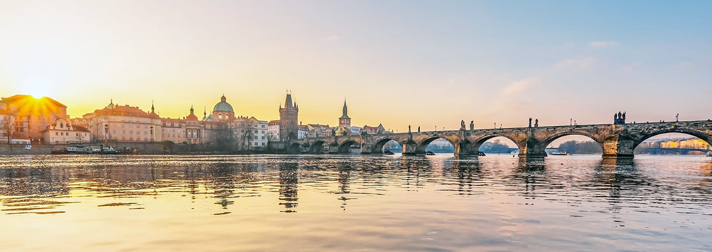 Historic stone bridge over a calm river at sunrise, with city skyline reflected in the water.