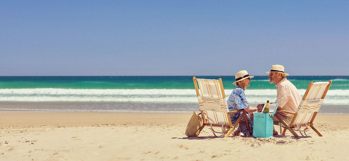 Two people sitting on deck chairs at a sandy beach with turquoise waves, enjoying drinks under a clear blue sky.