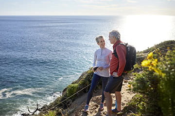 A mature couple take a break on their hike, they are standing by flowering plants on a cliff near the sea