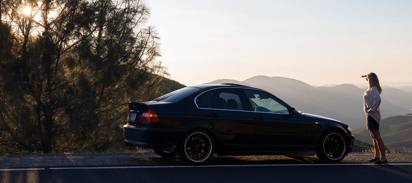 Woman stands beside parked car, looks across hills