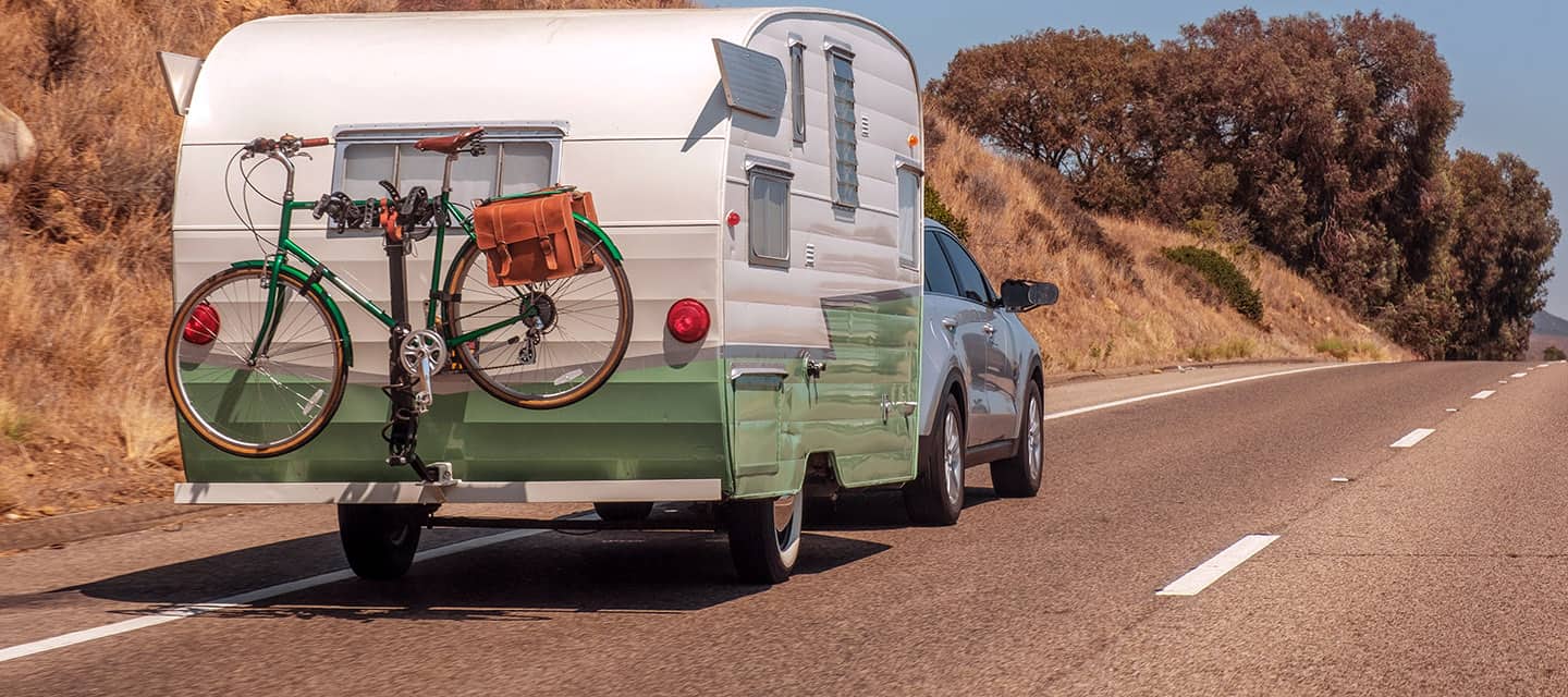 A retro caravan with a green bicycle on the back traveling on a country road.