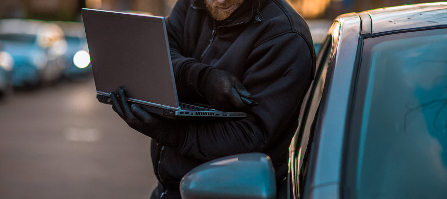 A keyless auto criminal dressed in black with tools of the trade hacking into the security of a car with a laptop in the early morning