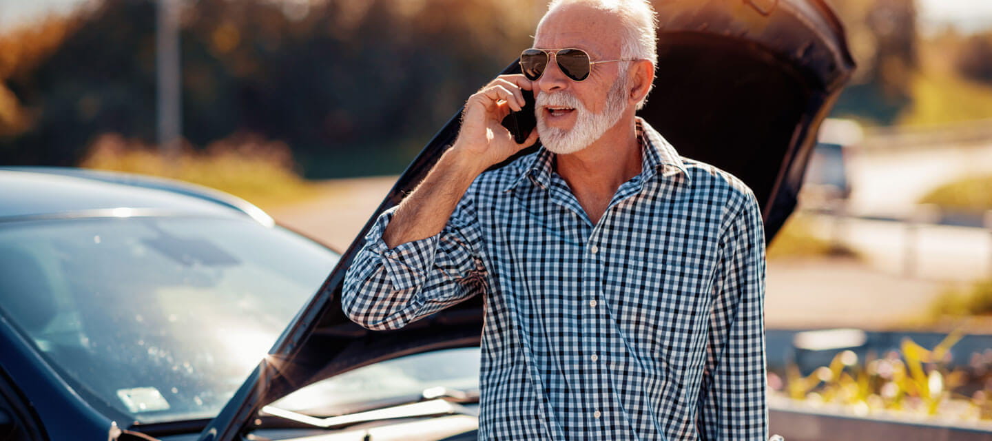 A senior man calling for assistance on his mobile phone next to a broken down car