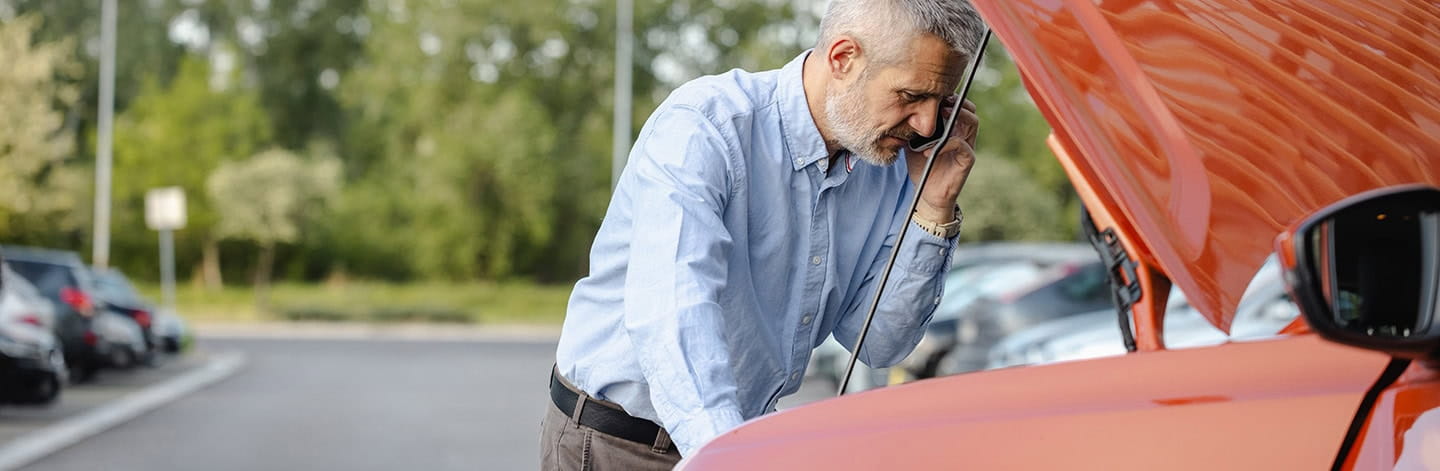 A mature man stands beside his broken-down car with the bonnet open, using his phone to call for assistance.