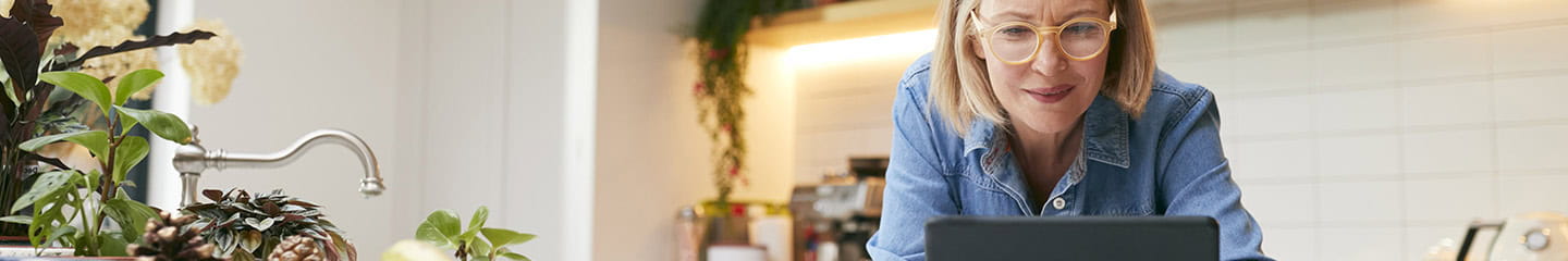 Mature woman at home in kitchen drinking coffee and looking at digital tablet