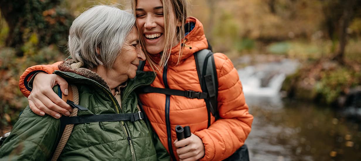 A senior woman and her adult daughter share a hub while hiking on an Autumn day.