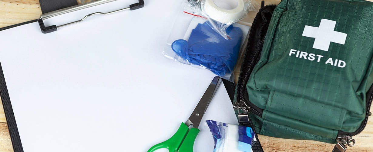 Green first aid kit on a wooden table with a clipboard and a pair of scissors and some dressings