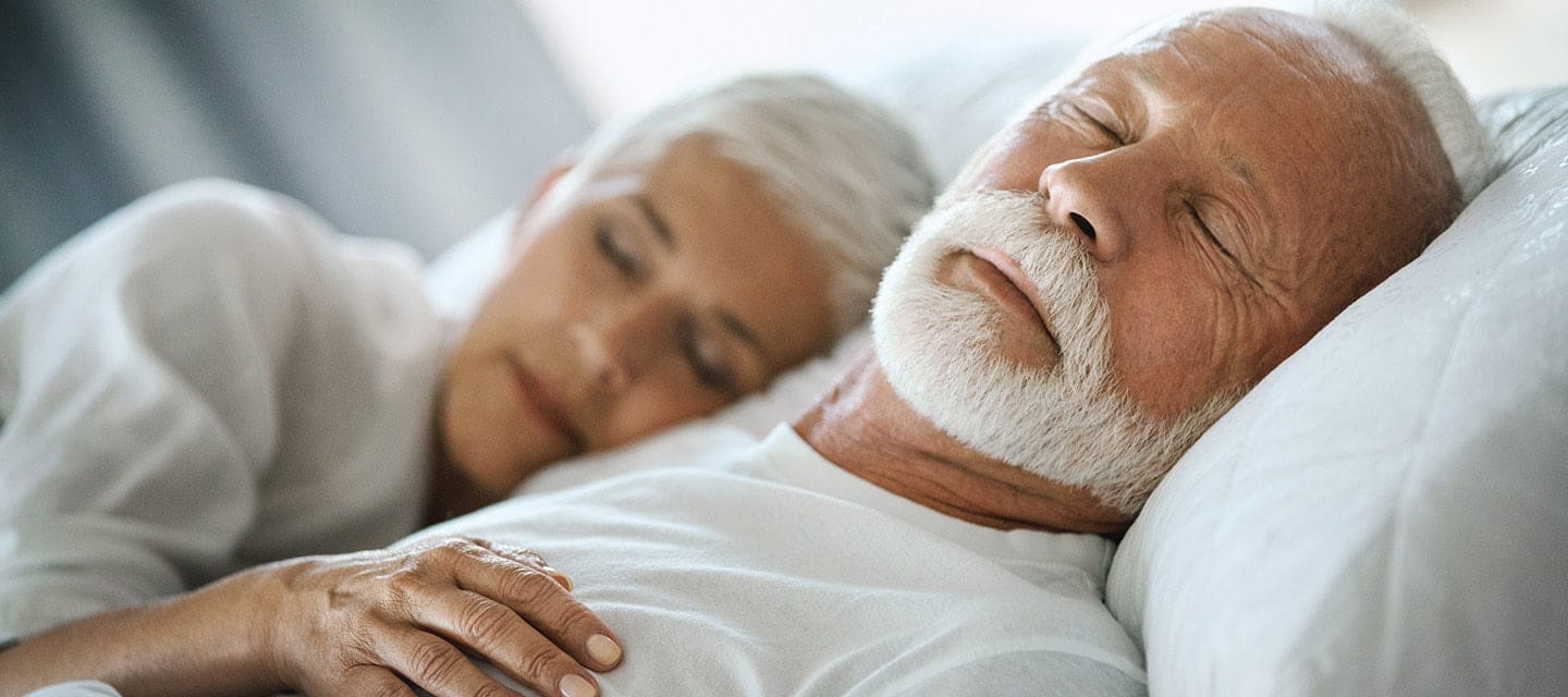 Closeup of mid 60's couple sleeping late in the morning in an already brightly lit bedroom.