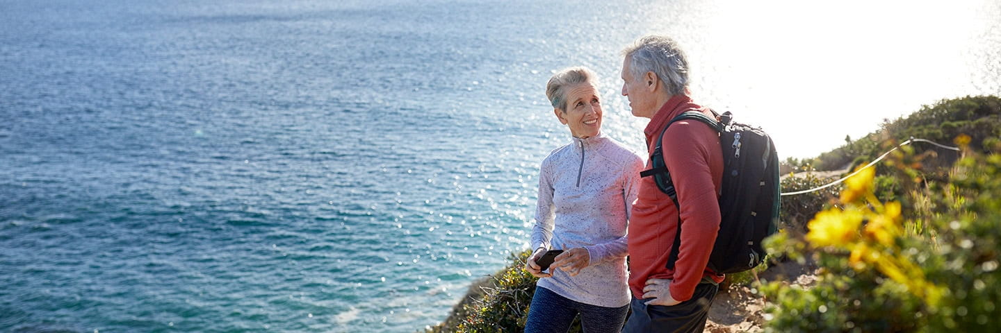 A mature couple take a break on their hike, they are standing by flowering plants on a cliff near the sea
