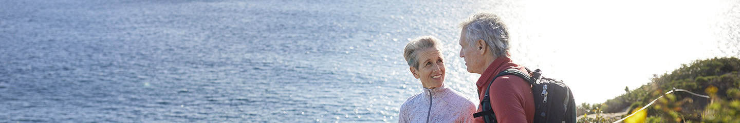 A mature couple take a break on their hike, they are standing by flowering plants on a cliff near the sea