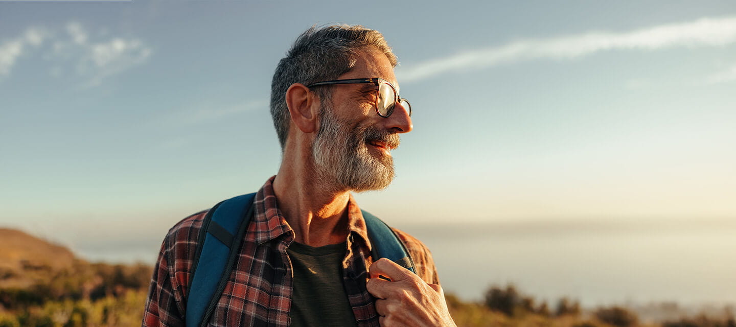 Happy mature hiker with a smile on his face while standing on top of a hill with a backpack.