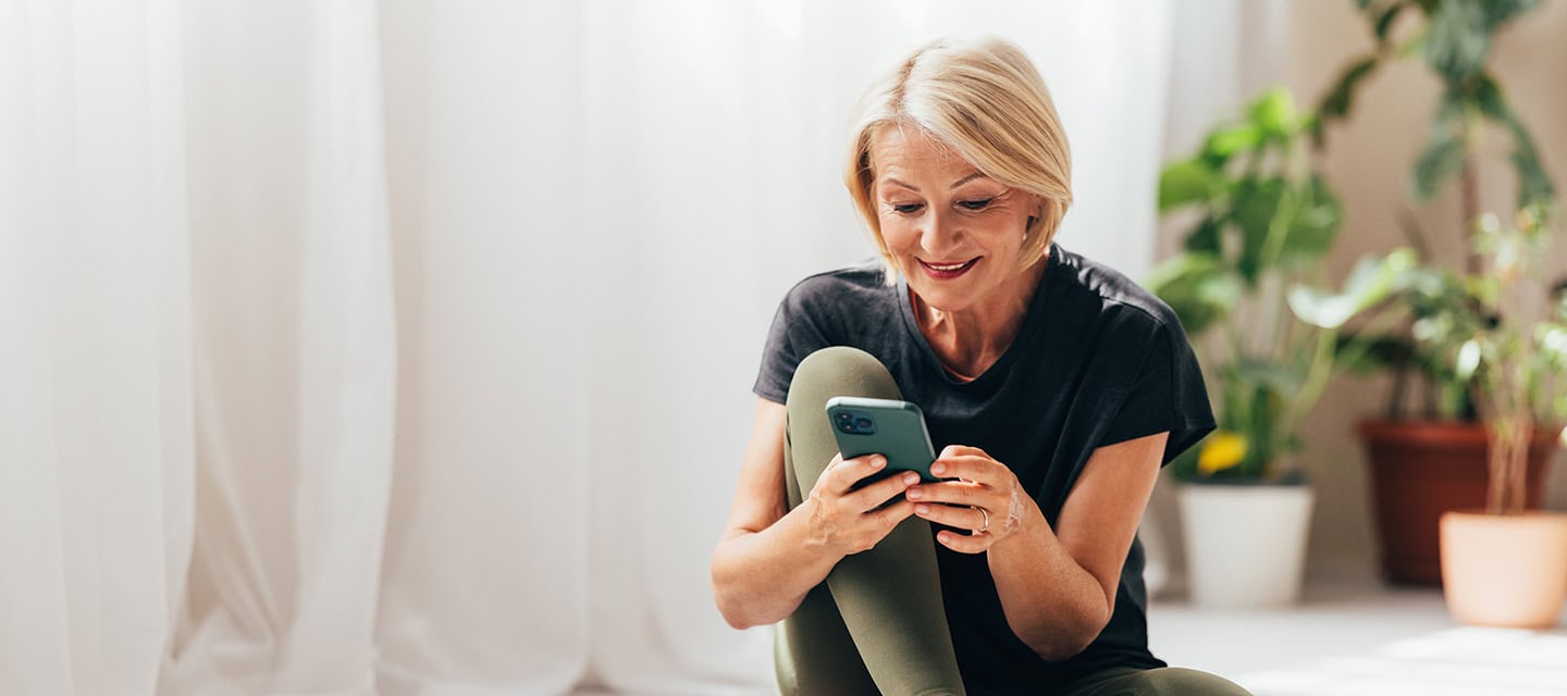 Mature woman texting on her smartphone after doing her daily workout at home on yoga mat.