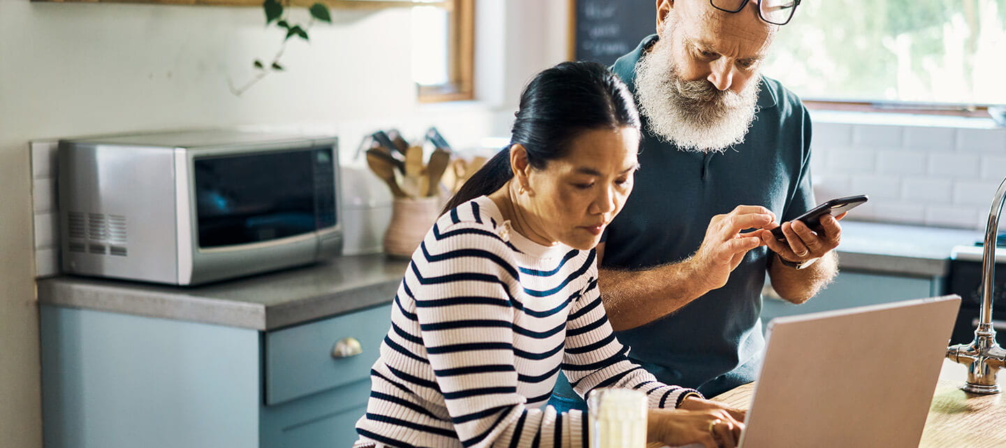 A mature couple using a laptop and phone in the kitchen at home.