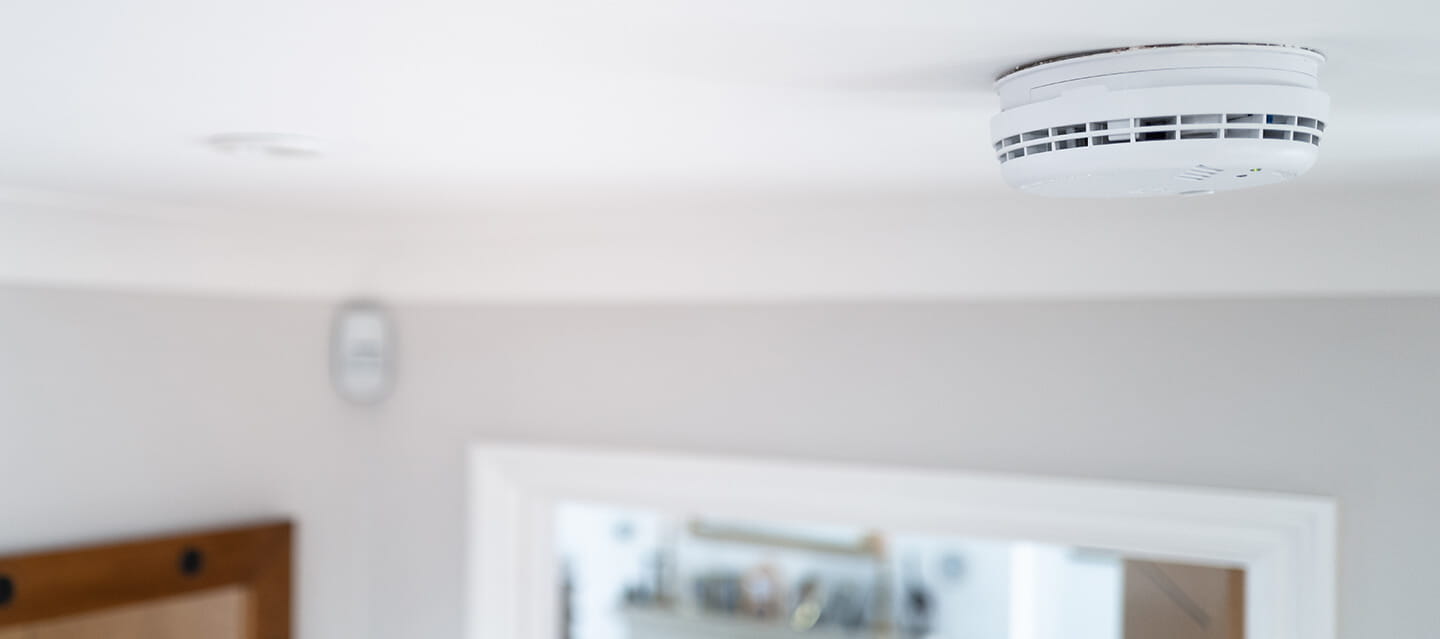 Shallow focus of a hard wired smoke detector seen in the downstairs hall, fixed to the ceiling. An open door leading to a living room can be seen.