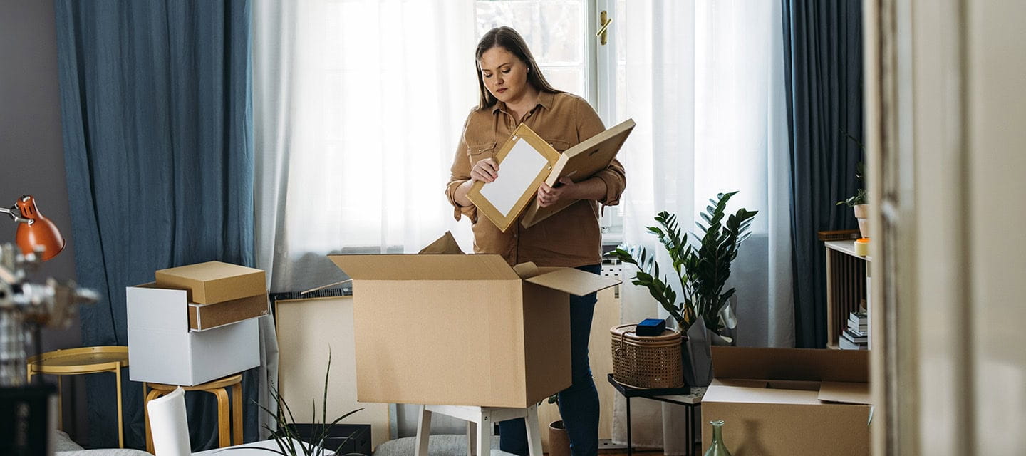 A woman in her 30's standing in the middle of a messy room, looking in a box in front of her and holding picture frames in her hands.