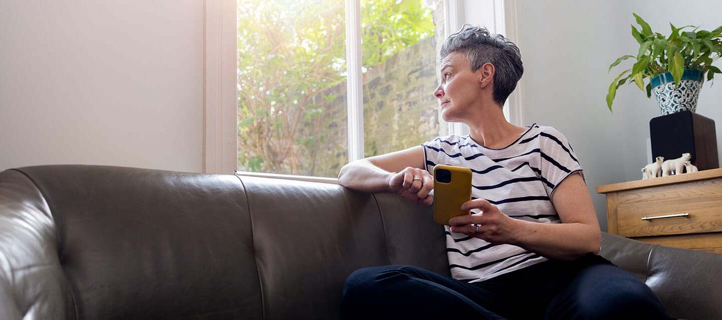 A mature female adult sitting on a leather sofa. She has her phone in her hands whilst looking out the window.